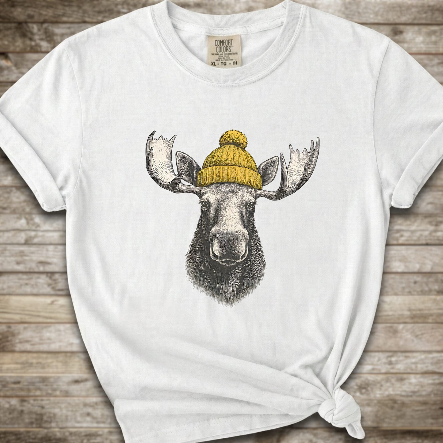 A white short-sleeve t-shirt with a graphic of a moose wearing a yellow knit beanie, displayed on a rustic wooden background.
