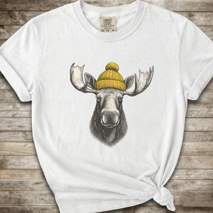 A white short-sleeve t-shirt with a graphic of a moose wearing a yellow knit beanie, displayed on a rustic wooden background.