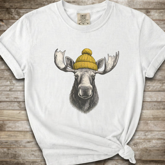 A white short-sleeve t-shirt with a graphic of a moose wearing a yellow knit beanie, displayed on a rustic wooden background.