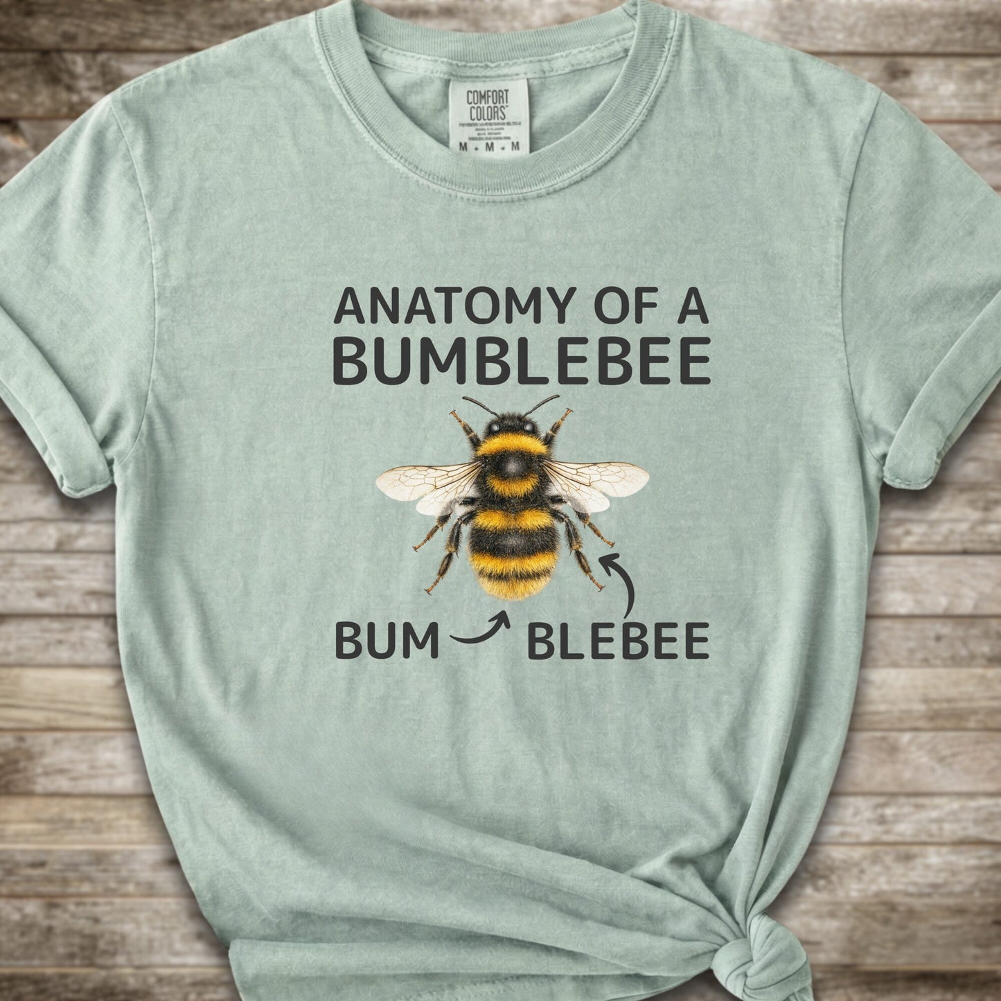 A light green graphic t-shirt with a knot at the bottom right, featuring a bumblebee illustration and the text 'ANATOMY OF A BUMBLEBEE' and 'BUM  BLEBEE'. The shirt is displayed flat on a rustic wooden background.