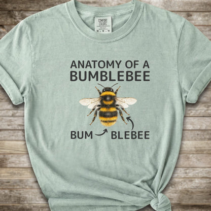 A light green graphic t-shirt with a knot at the bottom right, featuring a bumblebee illustration and the text 'ANATOMY OF A BUMBLEBEE' and 'BUM  BLEBEE'. The shirt is displayed flat on a rustic wooden background.