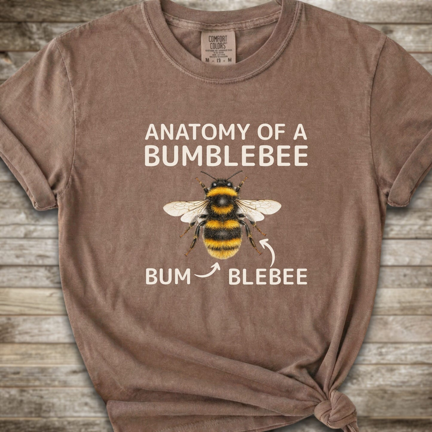 A brown graphic t-shirt with a knot at the bottom right, featuring a bumblebee illustration and the text 'ANATOMY OF A BUMBLEBEE' and 'BUM  BLEBEE'. The shirt is displayed flat on a rustic wooden background.