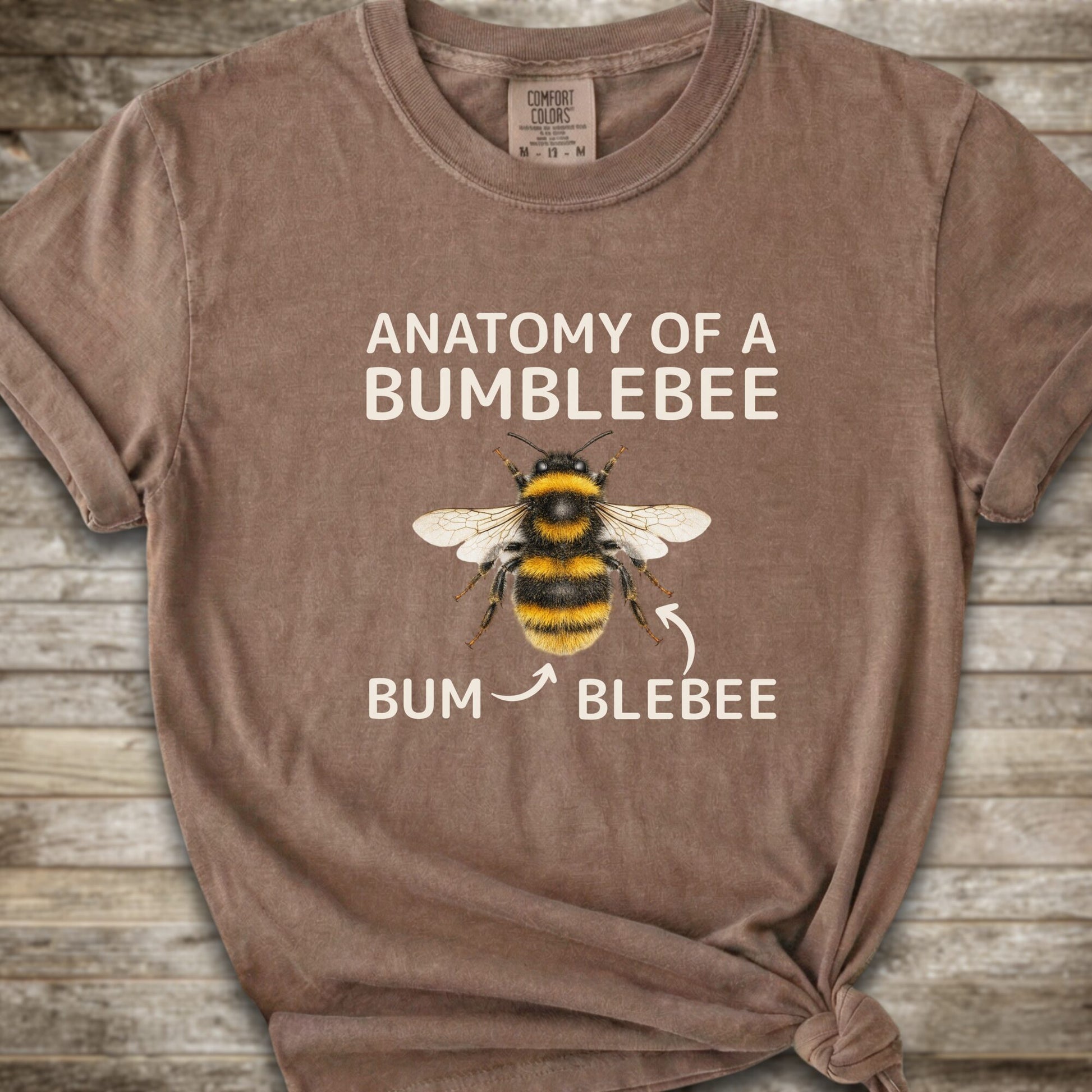 A brown graphic t-shirt with a knot at the bottom right, featuring a bumblebee illustration and the text 'ANATOMY OF A BUMBLEBEE' and 'BUM  BLEBEE'. The shirt is displayed flat on a rustic wooden background.