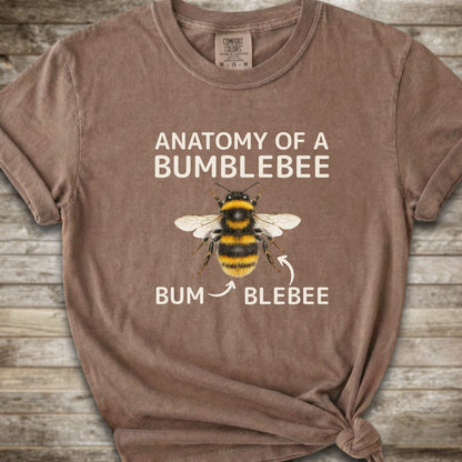 A brown graphic t-shirt with a knot at the bottom right, featuring a bumblebee illustration and the text 'ANATOMY OF A BUMBLEBEE' and 'BUM  BLEBEE'. The shirt is displayed flat on a rustic wooden background.