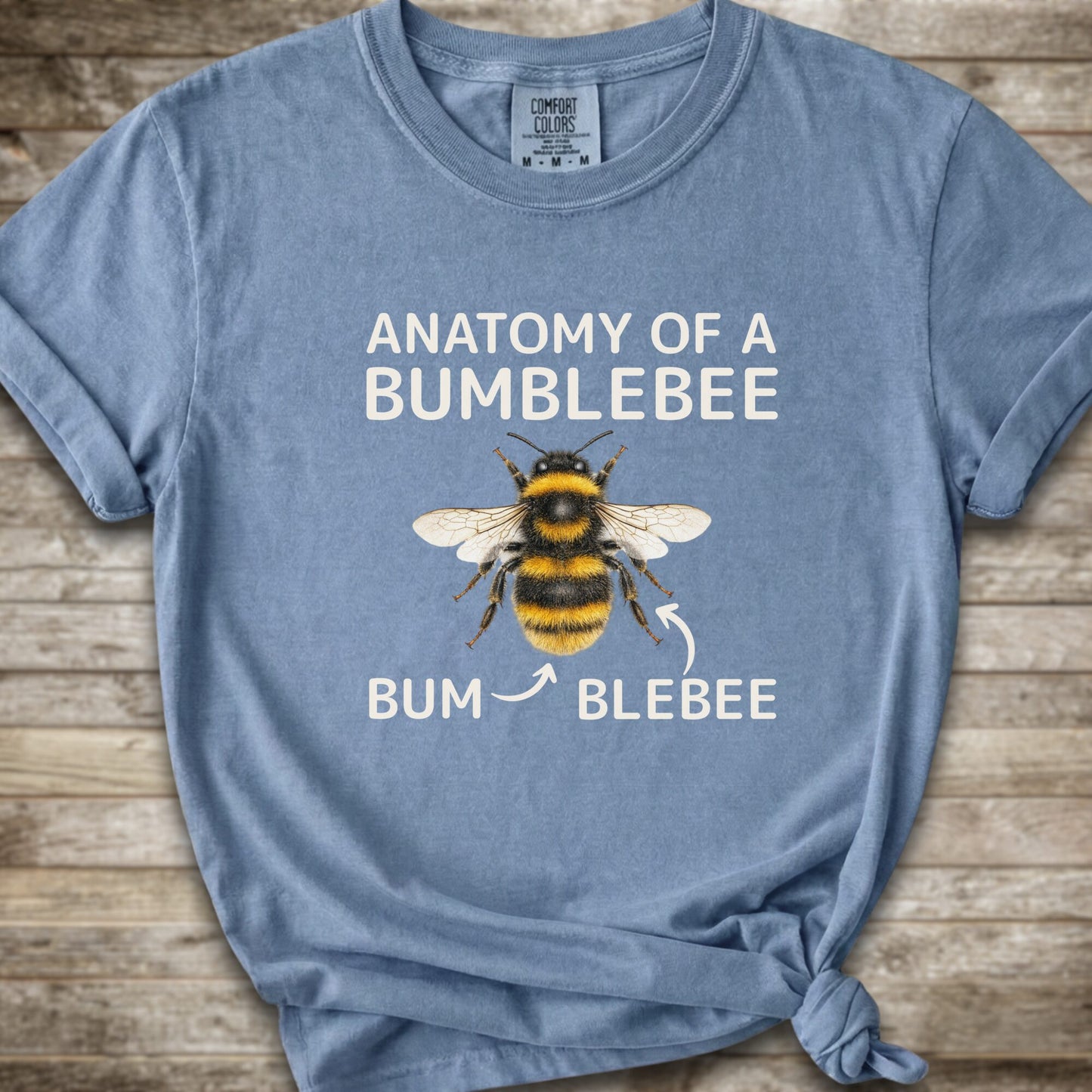 A light blue graphic t-shirt with a knot at the bottom right, featuring a bumblebee illustration and the text 'ANATOMY OF A BUMBLEBEE' and 'BUM  BLEBEE'. The shirt is displayed flat on a rustic wooden background.
