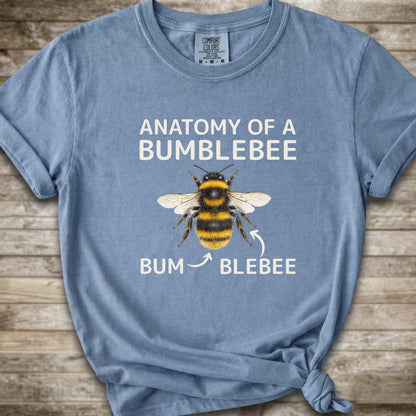 A light blue graphic t-shirt with a knot at the bottom right, featuring a bumblebee illustration and the text 'ANATOMY OF A BUMBLEBEE' and 'BUM  BLEBEE'. The shirt is displayed flat on a rustic wooden background.