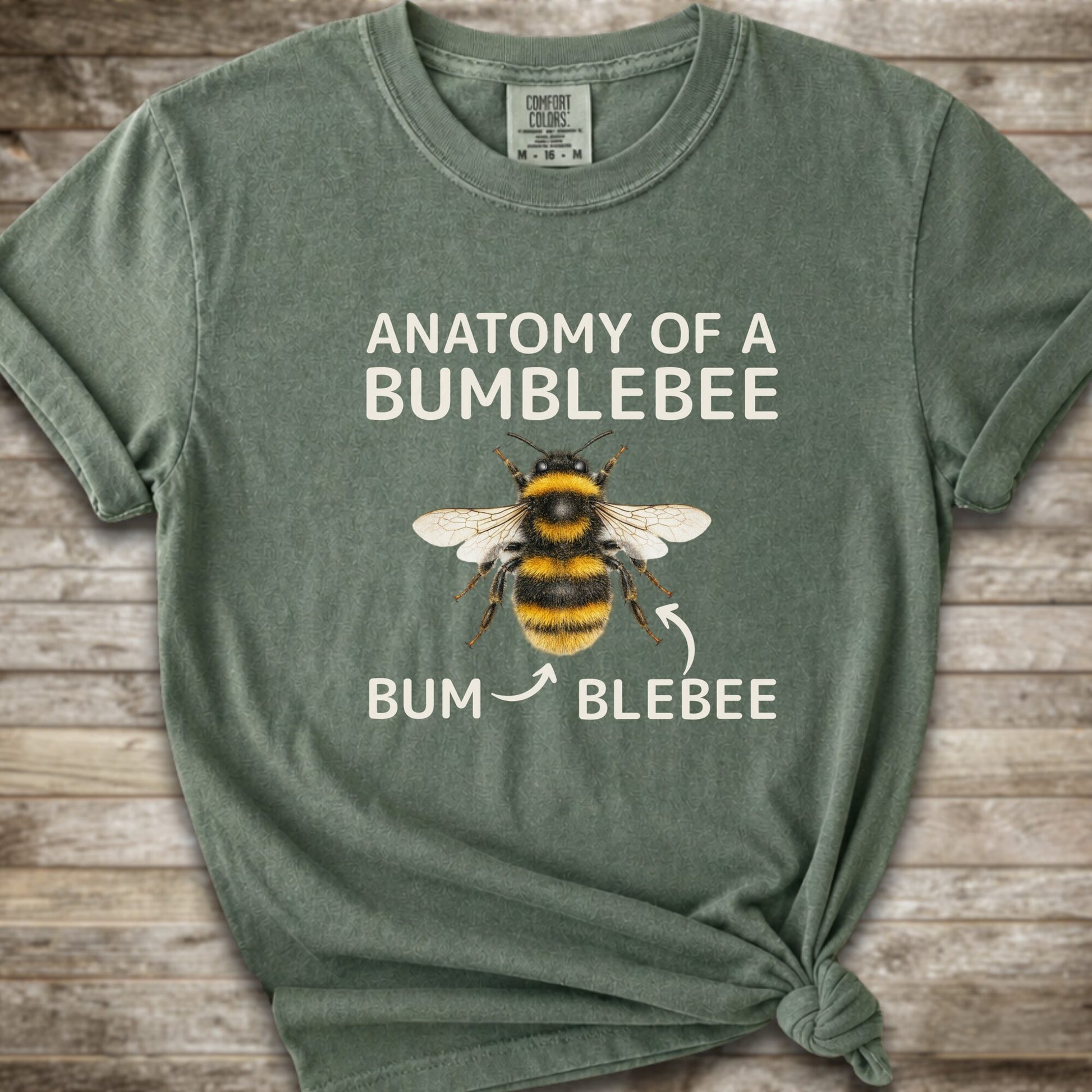 A moss green graphic t-shirt with a knot at the bottom right, featuring a bumblebee illustration and the text 'ANATOMY OF A BUMBLEBEE' and 'BUM  BLEBEE'. The shirt is displayed flat on a rustic wooden background.