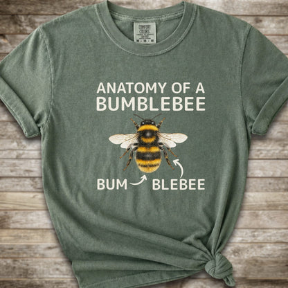 A moss green graphic t-shirt with a knot at the bottom right, featuring a bumblebee illustration and the text 'ANATOMY OF A BUMBLEBEE' and 'BUM  BLEBEE'. The shirt is displayed flat on a rustic wooden background.