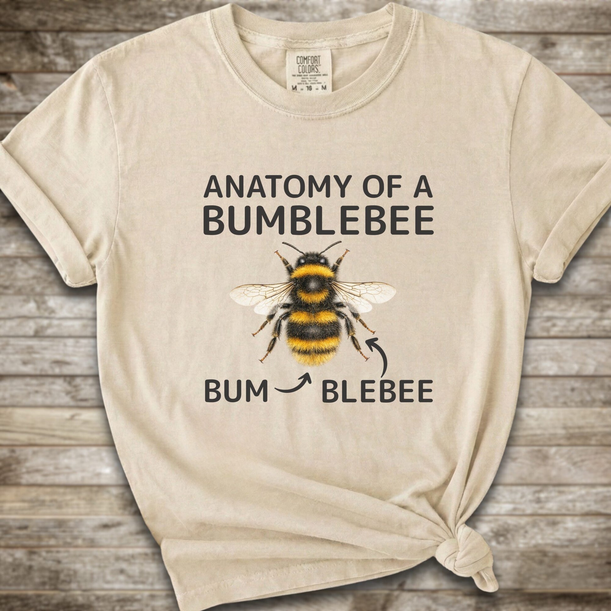 A Ivory graphic t-shirt with a knot at the bottom right, featuring a bumblebee illustration and the text 'ANATOMY OF A BUMBLEBEE' and 'BUM  BLEBEE'. The shirt is displayed flat on a rustic wooden background.