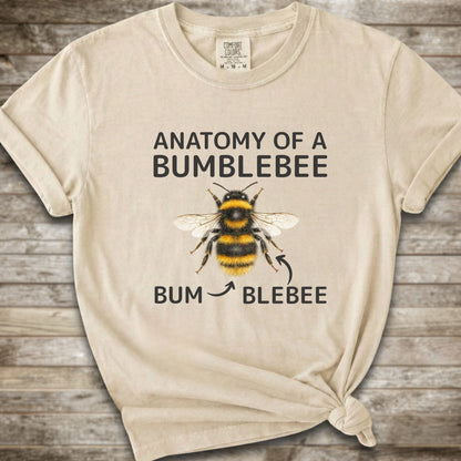 A Ivory graphic t-shirt with a knot at the bottom right, featuring a bumblebee illustration and the text 'ANATOMY OF A BUMBLEBEE' and 'BUM  BLEBEE'. The shirt is displayed flat on a rustic wooden background.