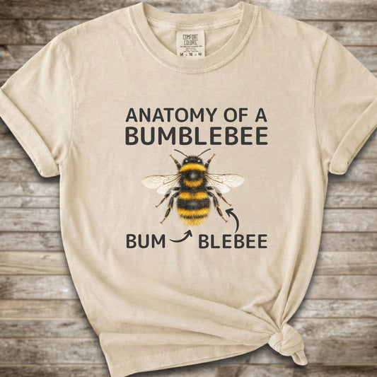 A Ivory graphic t-shirt with a knot at the bottom right, featuring a bumblebee illustration and the text 'ANATOMY OF A BUMBLEBEE' and 'BUM  BLEBEE'. The shirt is displayed flat on a rustic wooden background.