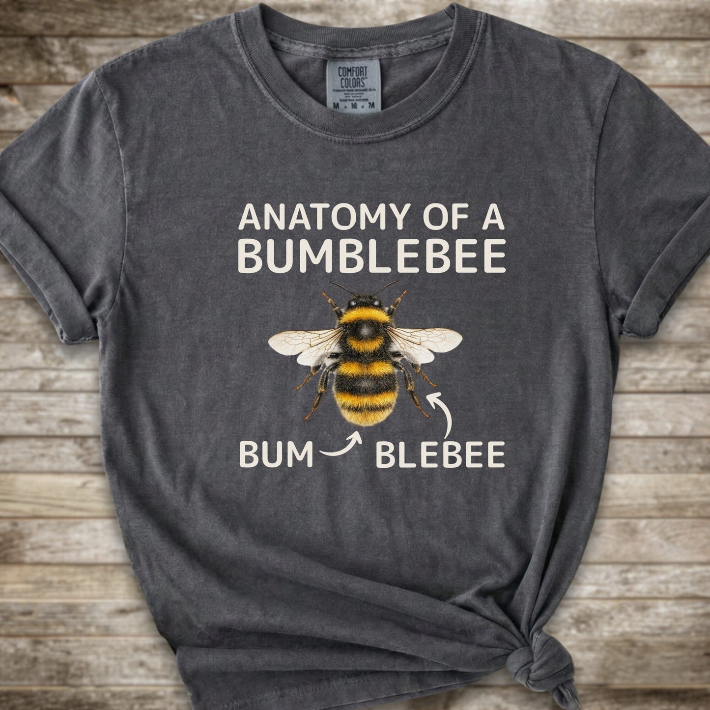 A dark grey graphic t-shirt with a knot at the bottom right, featuring a bumblebee illustration and the text 'ANATOMY OF A BUMBLEBEE' and 'BUM  BLEBEE'. The shirt is displayed flat on a rustic wooden background.