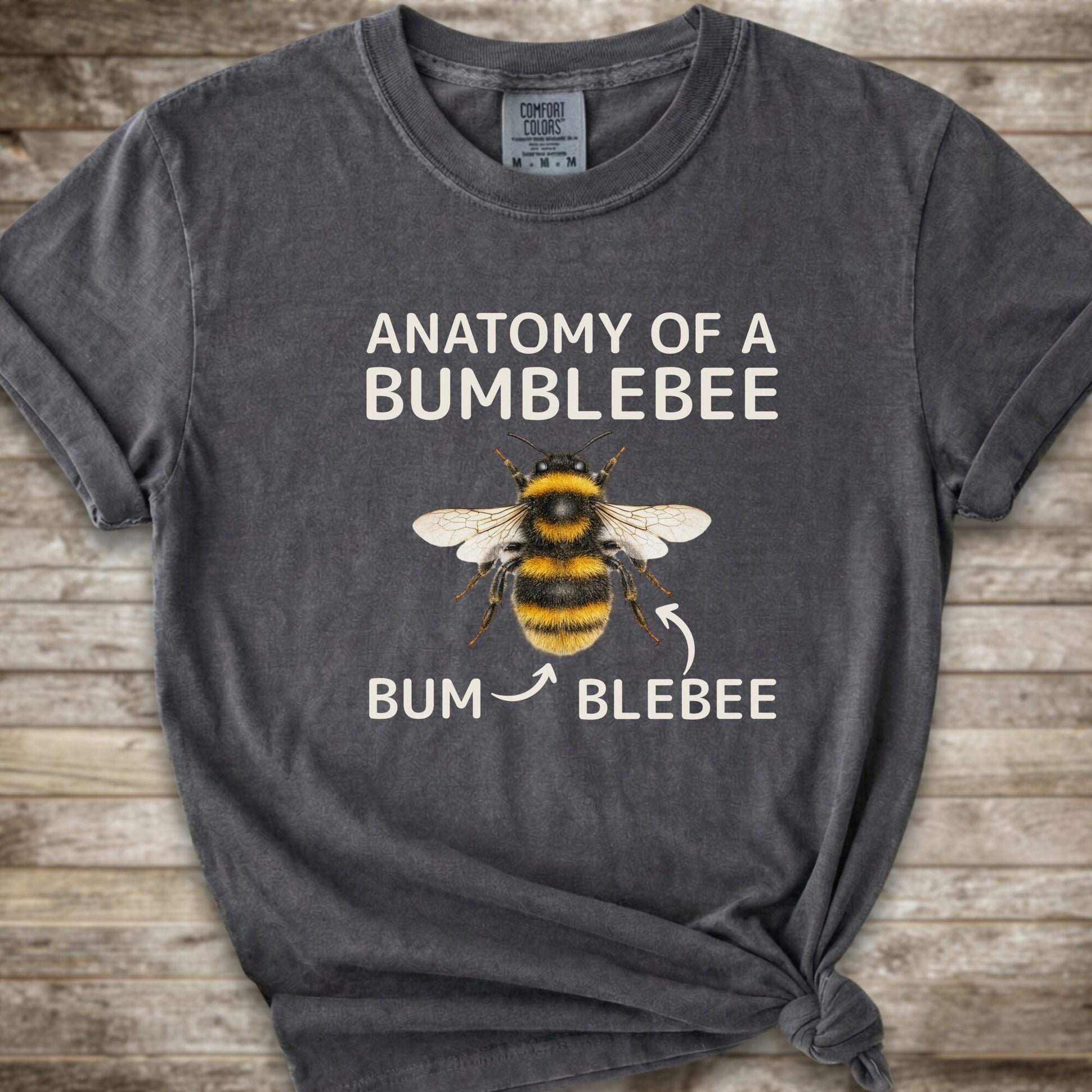 A dark grey graphic t-shirt with a knot at the bottom right, featuring a bumblebee illustration and the text 'ANATOMY OF A BUMBLEBEE' and 'BUM  BLEBEE'. The shirt is displayed flat on a rustic wooden background.