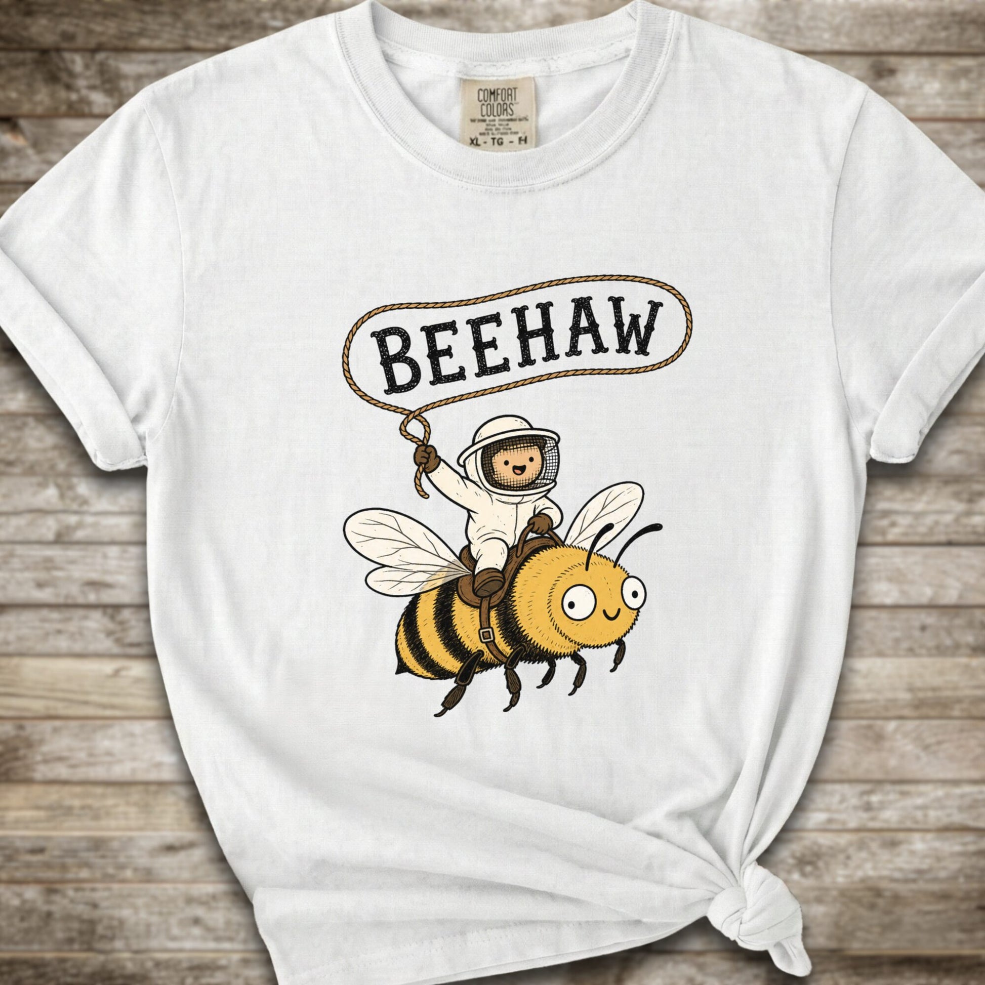 A white short-sleeve graphic T-shirt laid flat on a rustic wood background. The shirt has a printed design of a bee with a speech bubble above it containing the word “BEEHAW.