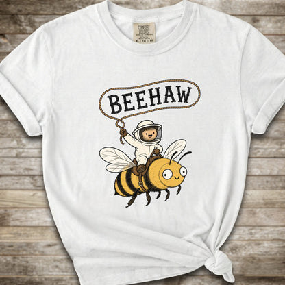 A white short-sleeve graphic T-shirt laid flat on a rustic wood background. The shirt has a printed design of a bee with a speech bubble above it containing the word “BEEHAW.