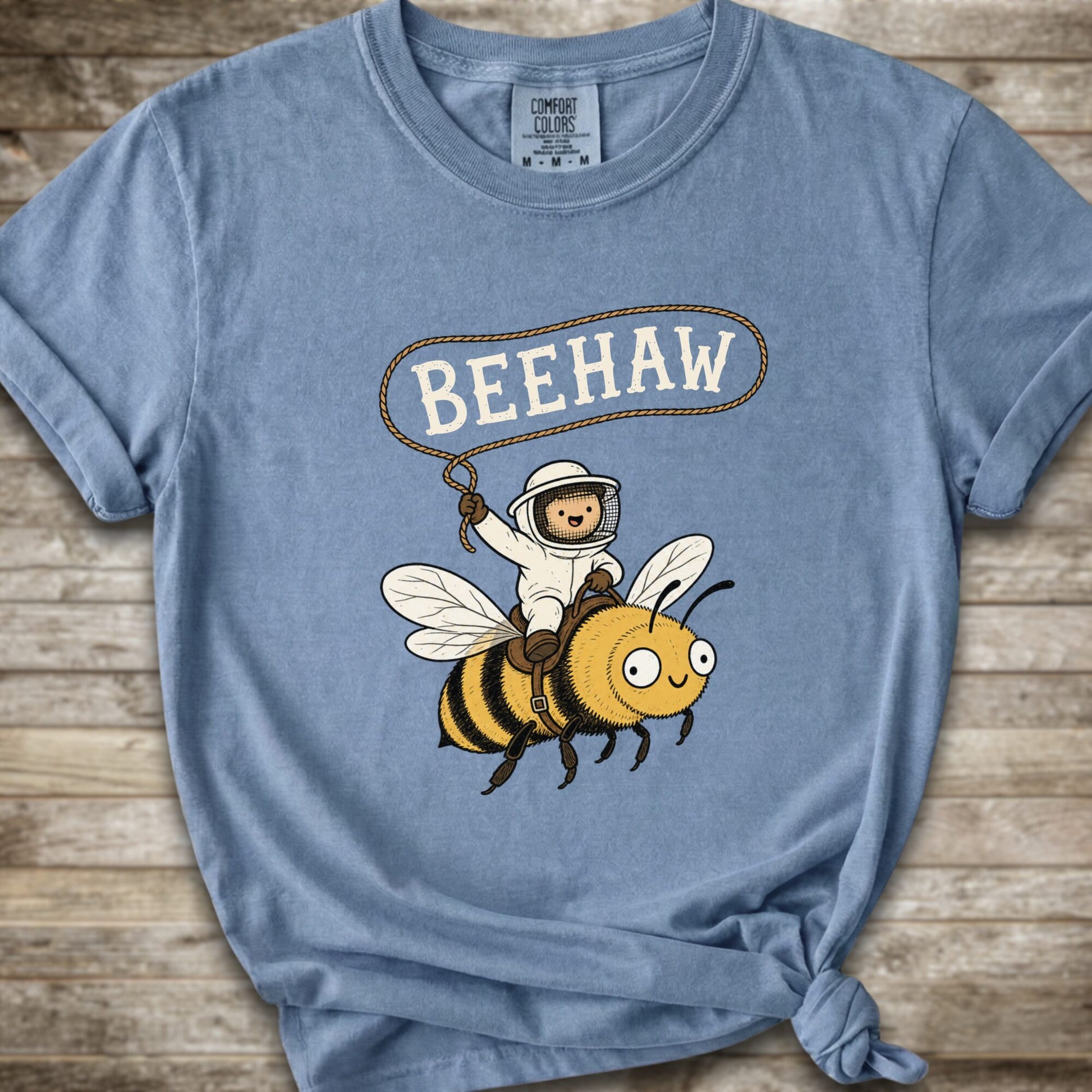 A light blue short-sleeve graphic T-shirt laid flat on a rustic wood background. The shirt has a printed design of a bee with a speech bubble above it containing the word “BEEHAW.