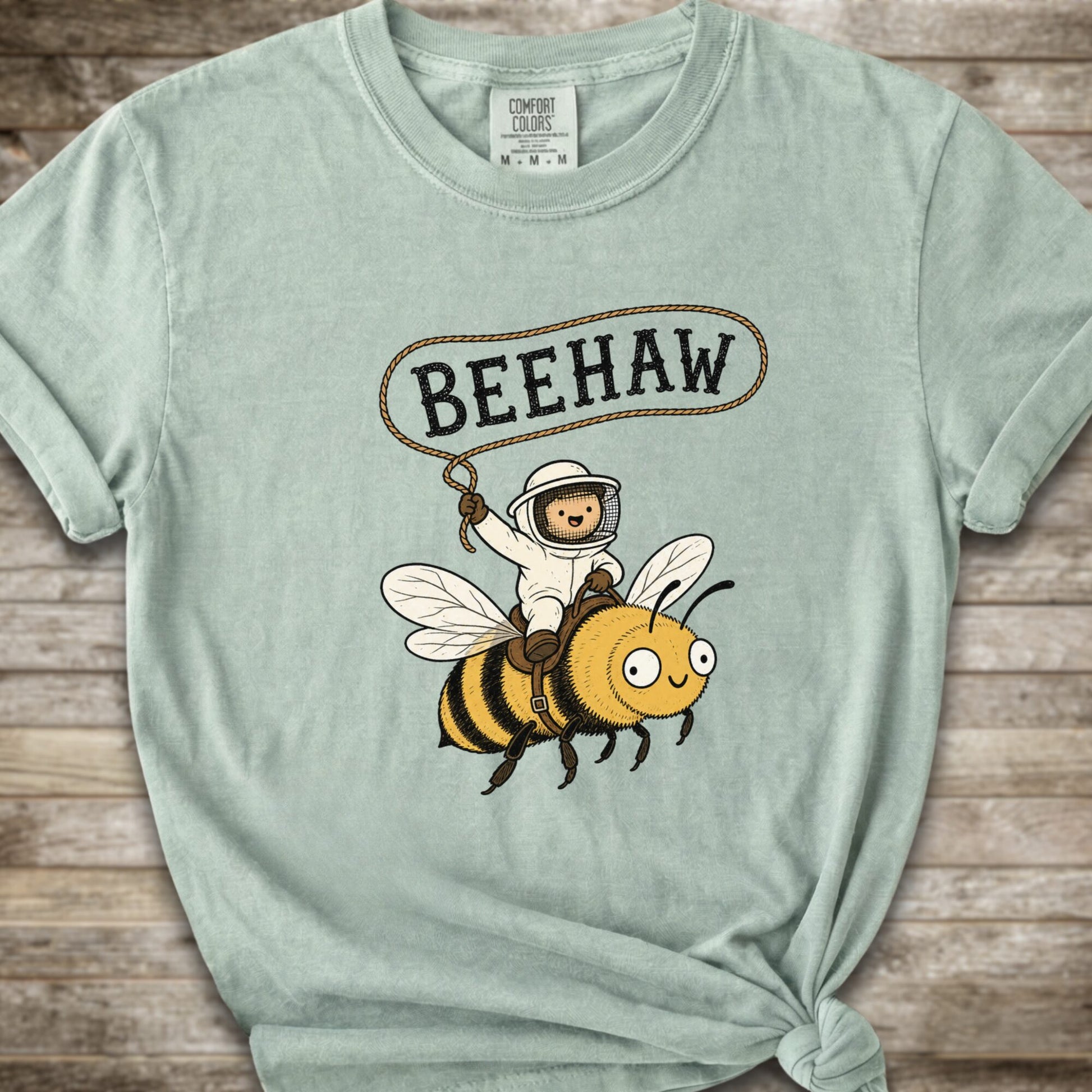 A light Green short-sleeve graphic T-shirt laid flat on a rustic wood background. The shirt has a printed design of a bee with a speech bubble above it containing the word “BEEHAW.
