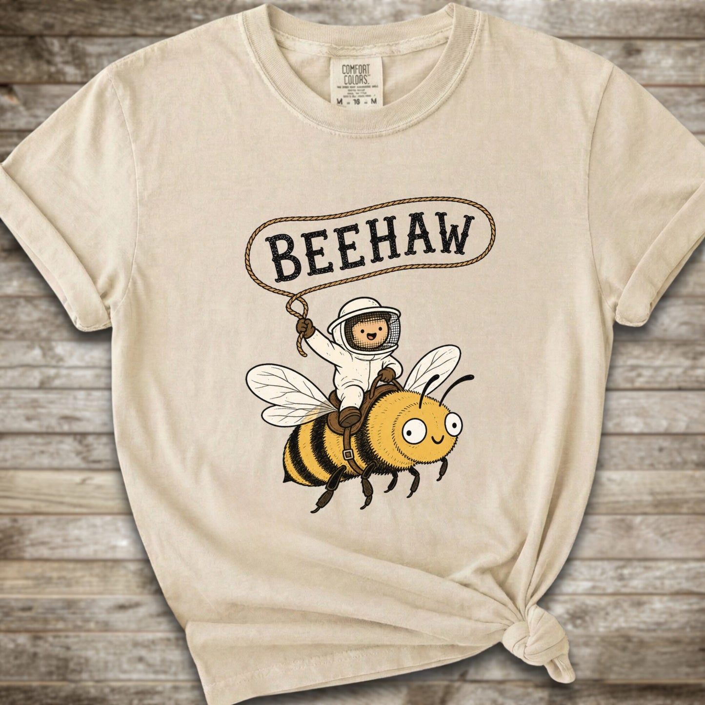 An ivory short-sleeve graphic T-shirt laid flat on a rustic wood background. The shirt has a printed design of a bee with a speech bubble above it containing the word “BEEHAW.