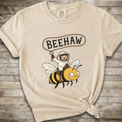 An ivory short-sleeve graphic T-shirt laid flat on a rustic wood background. The shirt has a printed design of a bee with a speech bubble above it containing the word “BEEHAW.