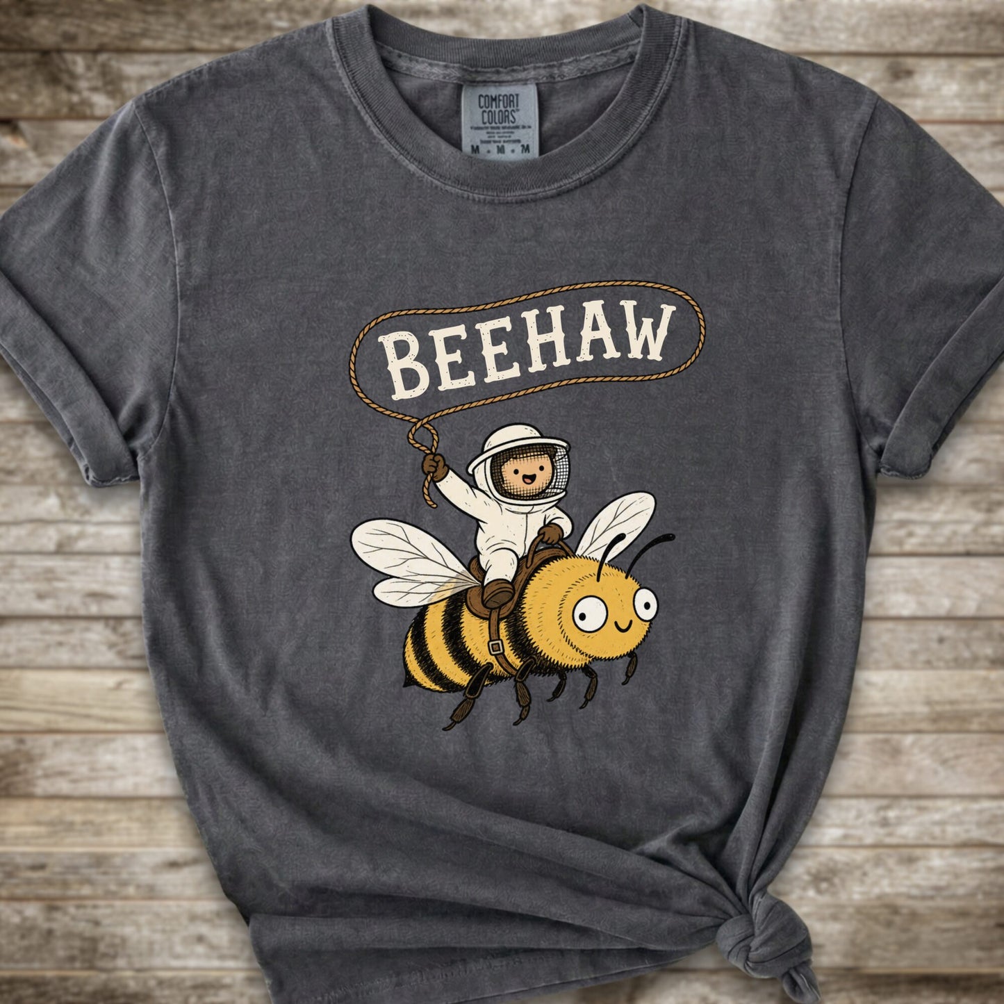 A dark grey short-sleeve graphic T-shirt laid flat on a rustic wood background. The shirt has a printed design of a bee with a speech bubble above it containing the word “BEEHAW.