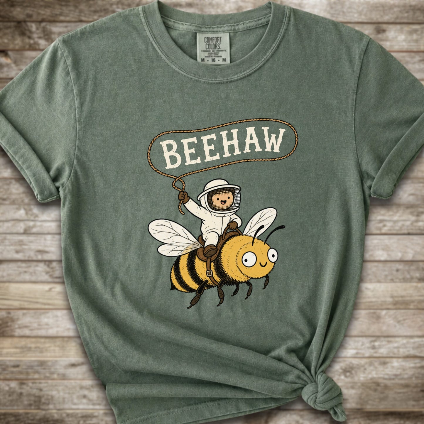 A moss green short-sleeve graphic T-shirt laid flat on a rustic wood background. The shirt has a printed design of a bee with a speech bubble above it containing the word “BEEHAW.