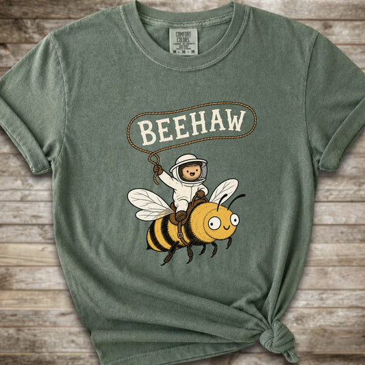 A moss green short-sleeve graphic T-shirt laid flat on a rustic wood background. The shirt has a printed design of a bee with a speech bubble above it containing the word “BEEHAW.