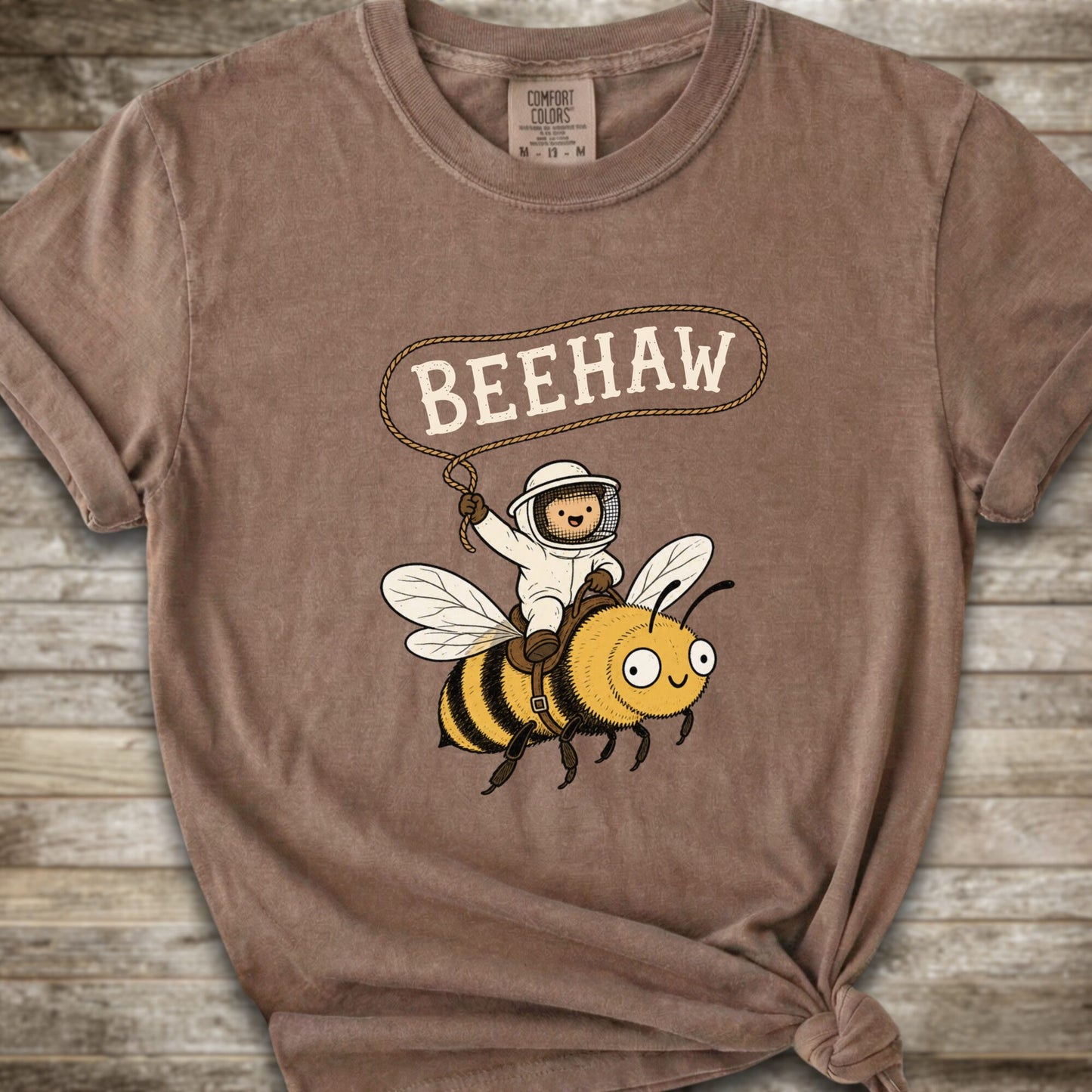 A brown short-sleeve graphic T-shirt laid flat on a rustic wood background. The shirt has a printed design of a bee with a speech bubble above it containing the word “BEEHAW.