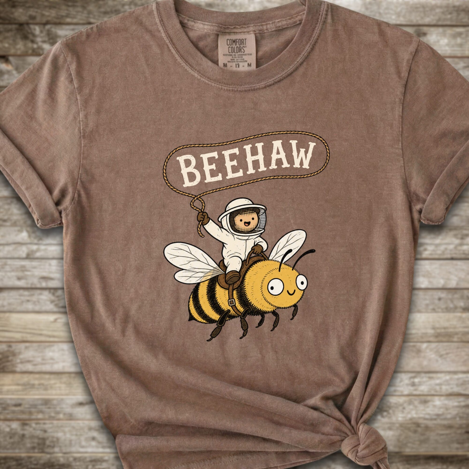 A brown short-sleeve graphic T-shirt laid flat on a rustic wood background. The shirt has a printed design of a bee with a speech bubble above it containing the word “BEEHAW.