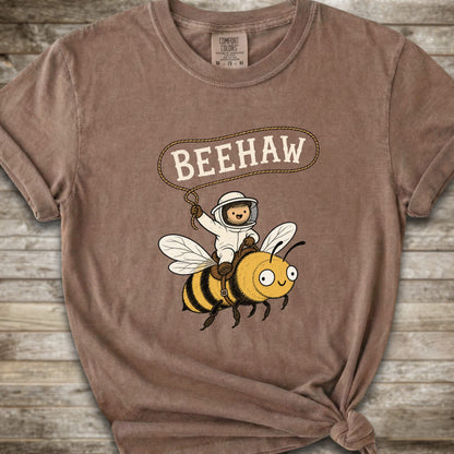 A brown short-sleeve graphic T-shirt laid flat on a rustic wood background. The shirt has a printed design of a bee with a speech bubble above it containing the word “BEEHAW.