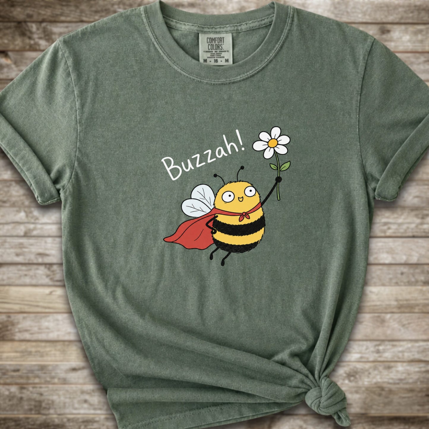A moss green Comfort Colors t-shirt with a cartoon bee graphic holding a white flower and the text 'Buzzah!' printed on the front, styled on a rustic wooden background.