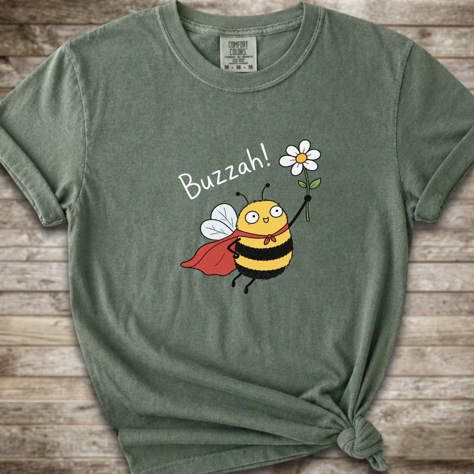 A moss green Comfort Colors t-shirt with a cartoon bee graphic holding a white flower and the text 'Buzzah!' printed on the front, styled on a rustic wooden background.