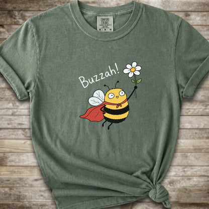 A moss green Comfort Colors t-shirt with a cartoon bee graphic holding a white flower and the text 'Buzzah!' printed on the front, styled on a rustic wooden background.