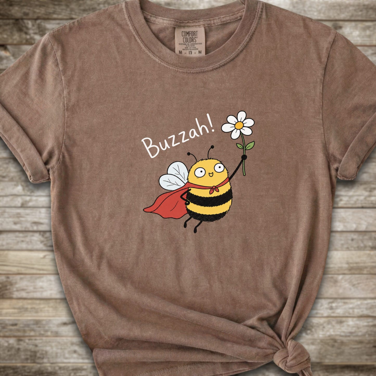 A brown Comfort Colors t-shirt with a cartoon bee graphic holding a white flower and the text 'Buzzah!' printed on the front, styled on a rustic wooden background.