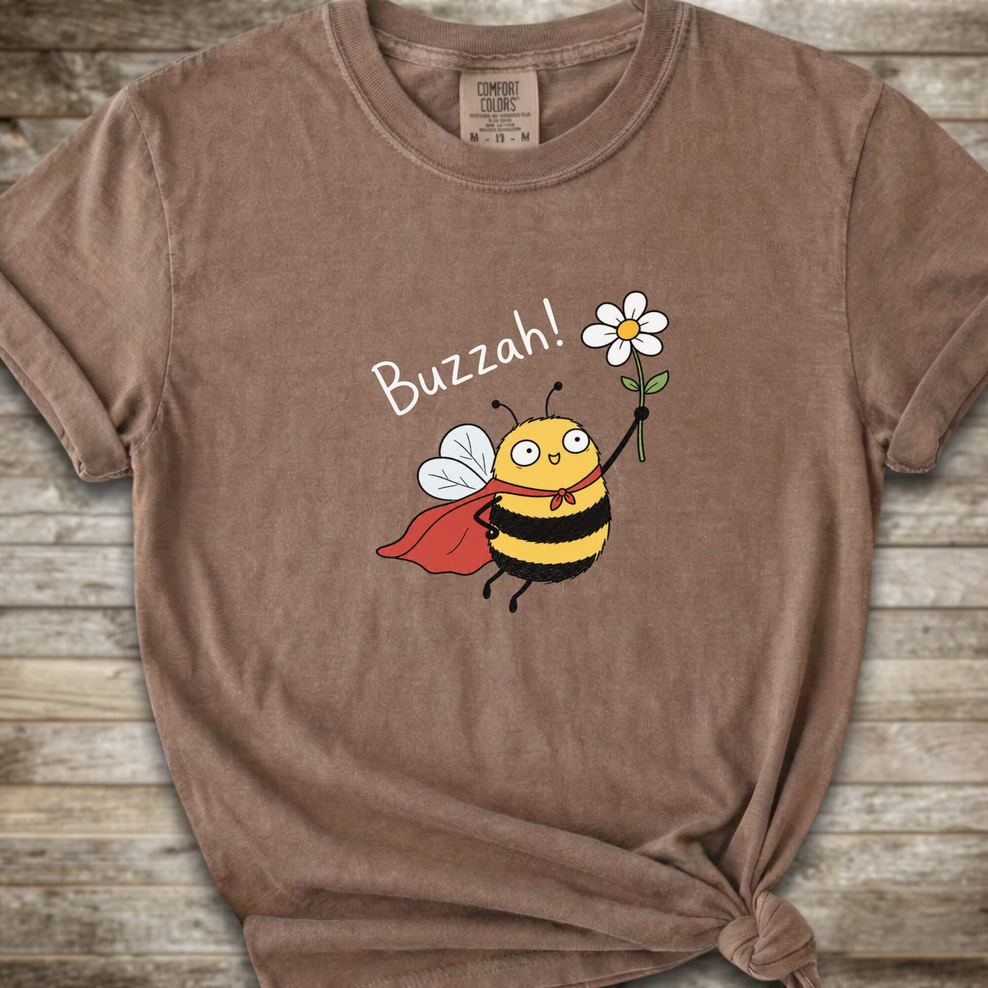 A brown Comfort Colors t-shirt with a cartoon bee graphic holding a white flower and the text 'Buzzah!' printed on the front, styled on a rustic wooden background.