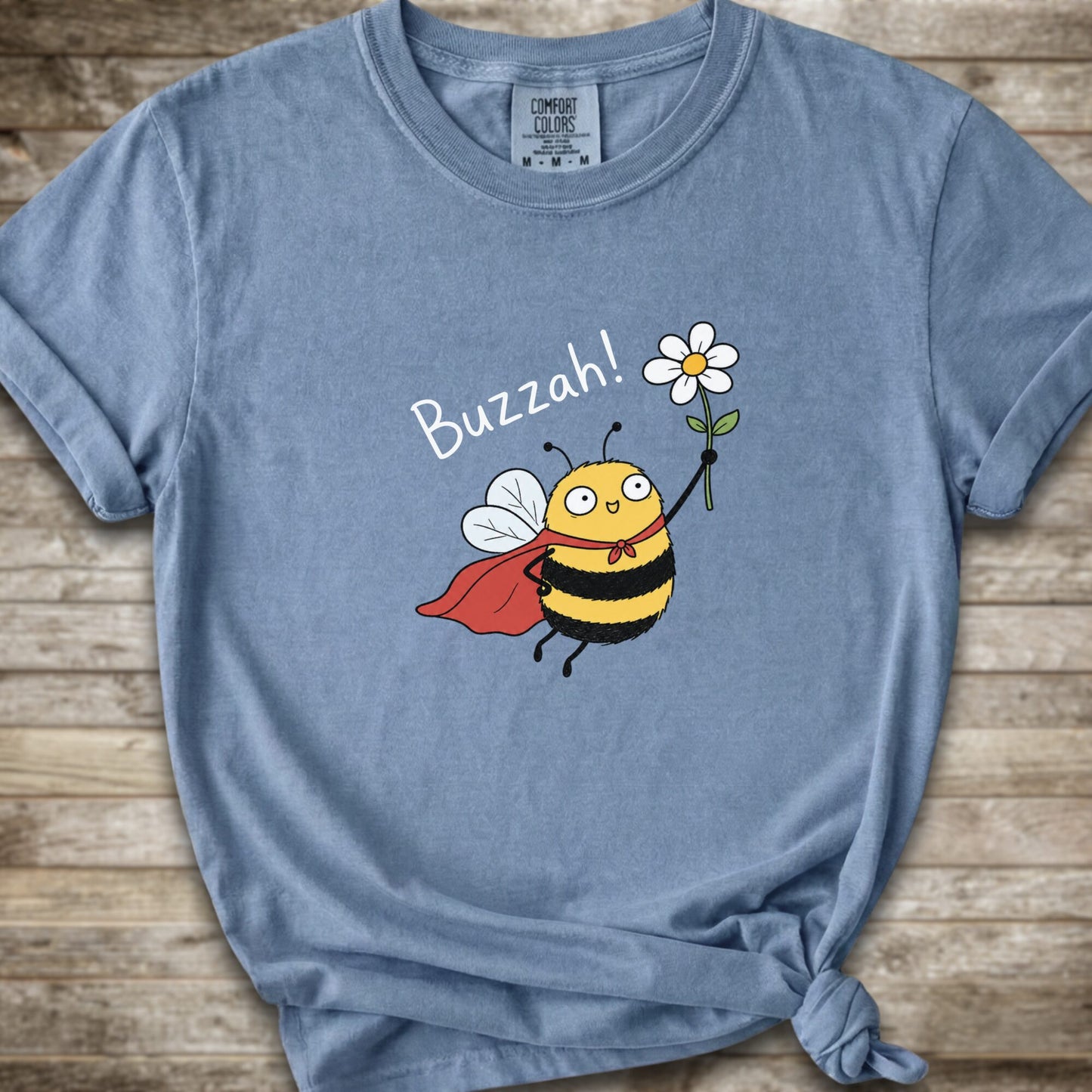 A light blue Comfort Colors t-shirt with a cartoon bee graphic holding a white flower and the text 'Buzzah!' printed on the front, styled on a rustic wooden background.
