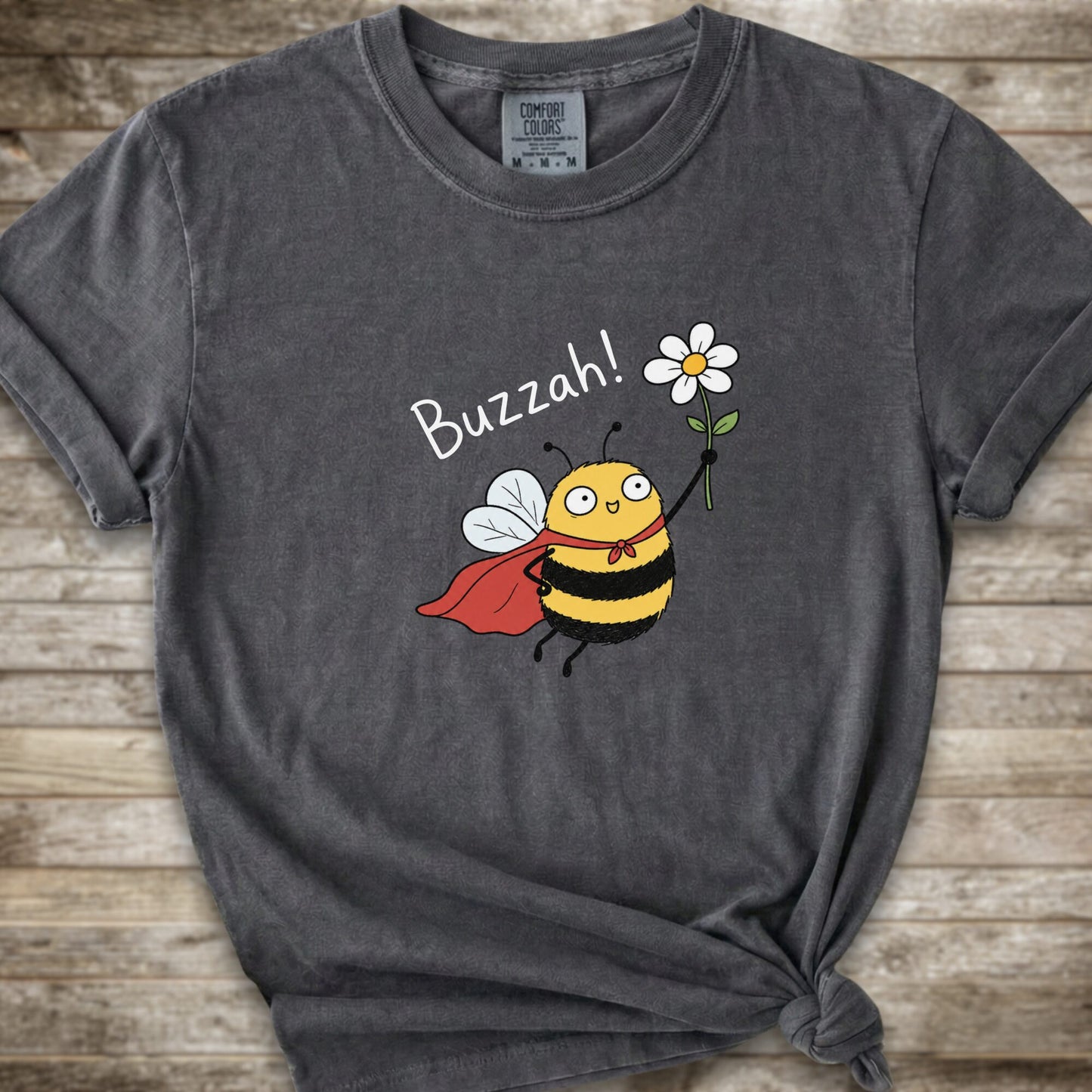 A dark grey Comfort Colors t-shirt with a cartoon bee graphic holding a white flower and the text 'Buzzah!' printed on the front, styled on a rustic wooden background.