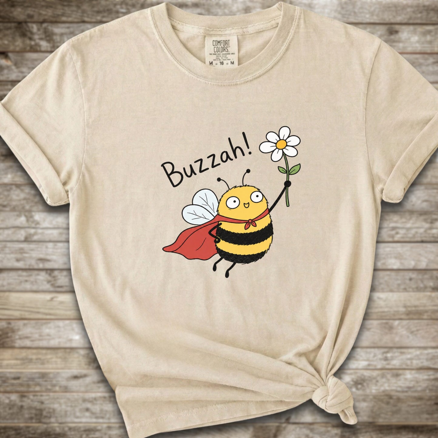 An Ivory Comfort Colors t-shirt with a cartoon bee graphic holding a white flower and the text 'Buzzah!' printed on the front, styled on a rustic wooden background.