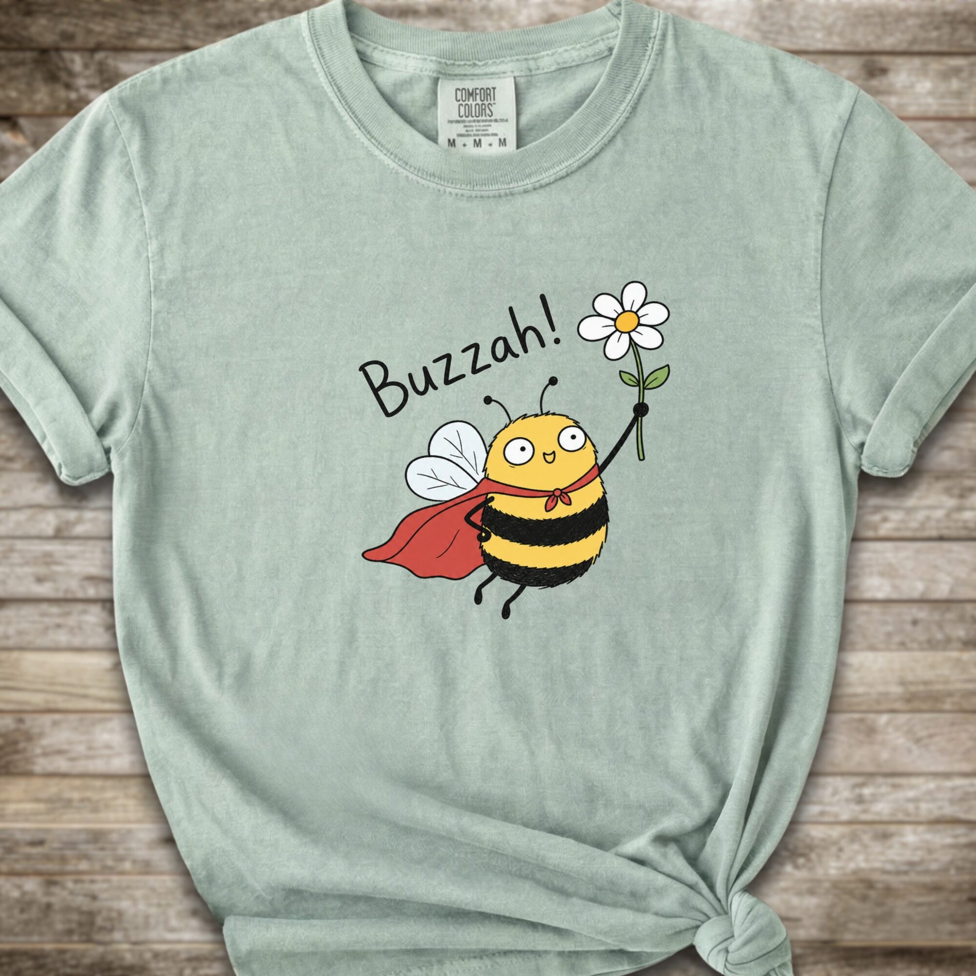 A light green Comfort Colors t-shirt with a cartoon bee graphic holding a white flower and the text 'Buzzah!' printed on the front, styled on a rustic wooden background.