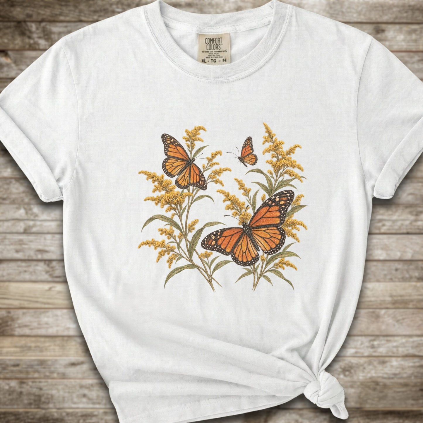 A white short-sleeve t-shirt with a graphic print of orange monarch butterflies and yellow Goldenrod, styled with a knot at the bottom hem against a rustic wooden background.
