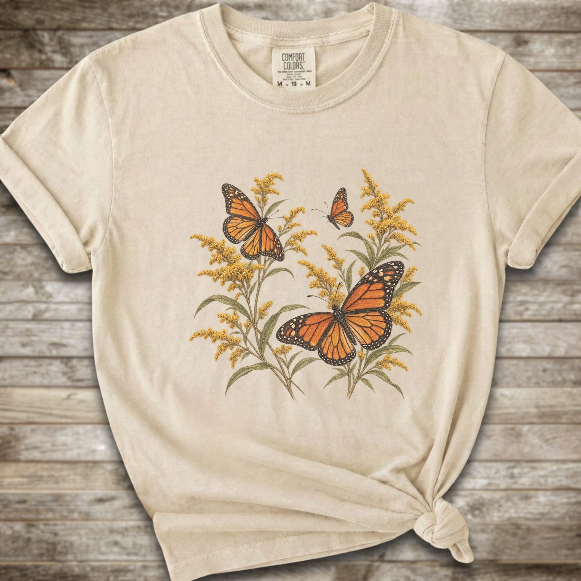 An Ivory short-sleeve t-shirt with a graphic print of orange monarch butterflies and yellow Goldenrod, styled with a knot at the bottom hem against a rustic wooden background.