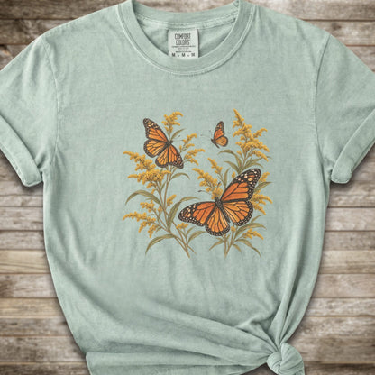 A light green short-sleeve t-shirt with a graphic print of orange monarch butterflies and yellow Goldenrod, styled with a knot at the bottom hem against a rustic wooden background.