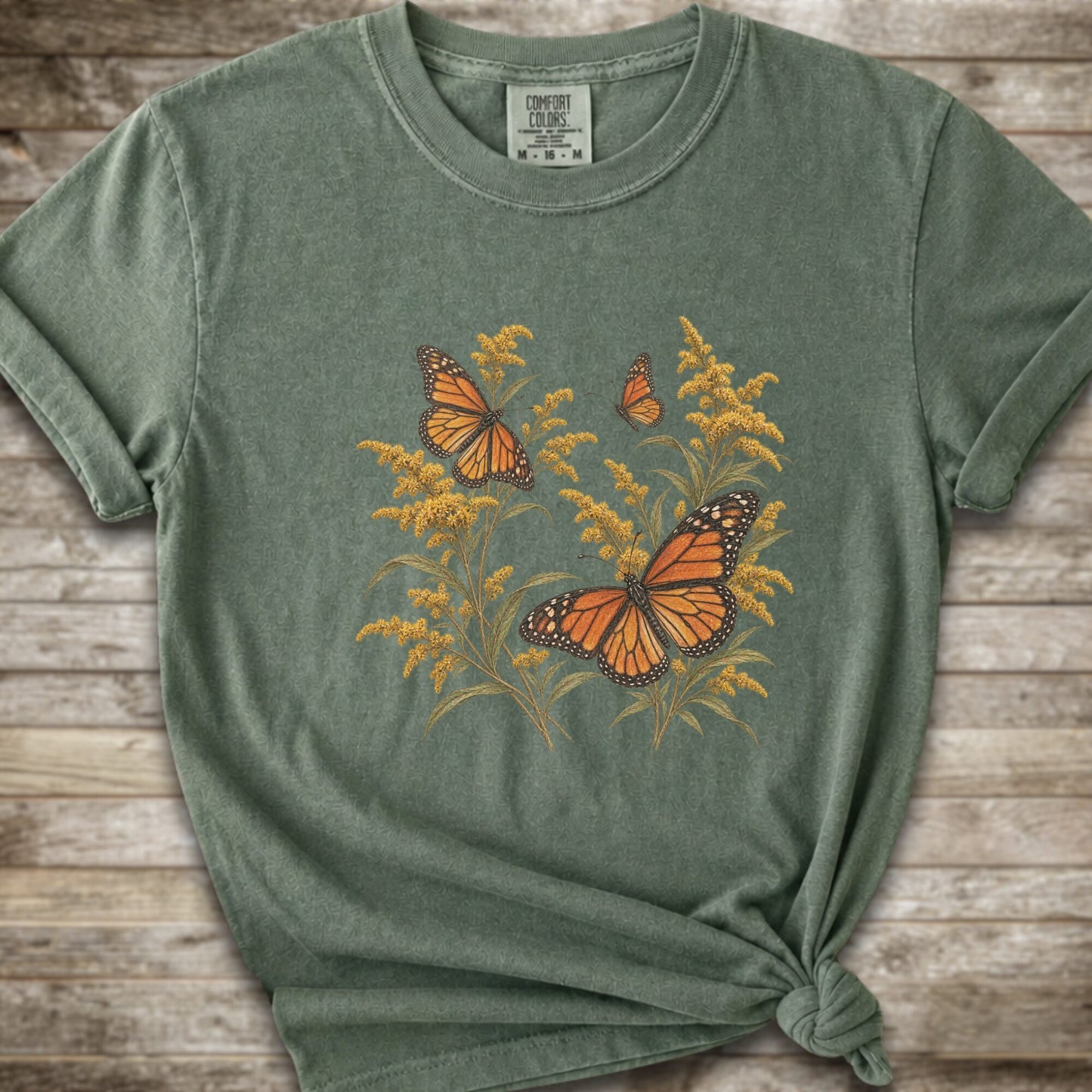 A moss green short-sleeve t-shirt with a graphic print of orange monarch butterflies and yellow Goldenrod, styled with a knot at the bottom hem against a rustic wooden background.