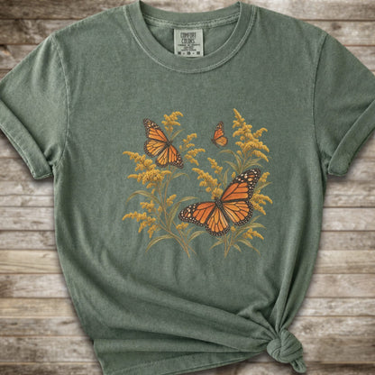 A moss green short-sleeve t-shirt with a graphic print of orange monarch butterflies and yellow Goldenrod, styled with a knot at the bottom hem against a rustic wooden background.