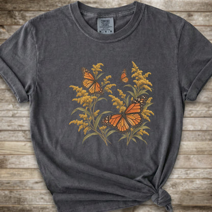 A dark grey short-sleeve t-shirt with a graphic print of orange monarch butterflies and yellow Goldenrod, styled with a knot at the bottom hem against a rustic wooden background.