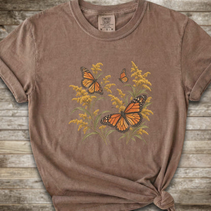 A brown short-sleeve t-shirt with a graphic print of orange monarch butterflies and yellow Goldenrod, styled with a knot at the bottom hem against a rustic wooden background.
