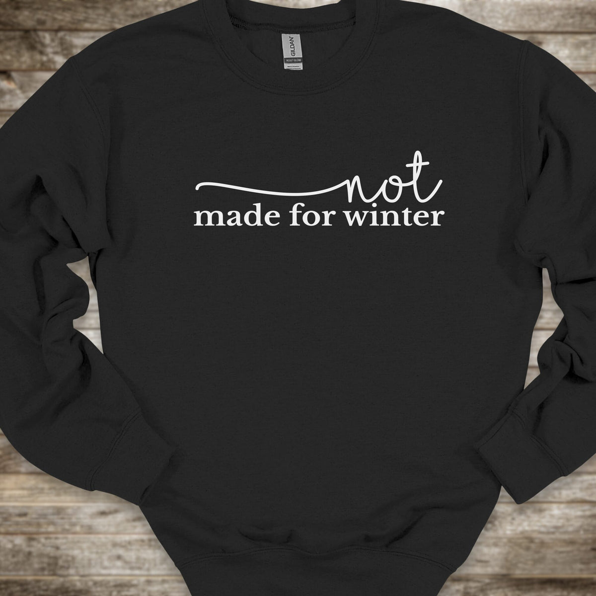 A black crewneck sweatshirt with the phrase 'not made for winter' printed in white across the chest, laid flat on a rustic wooden surface.