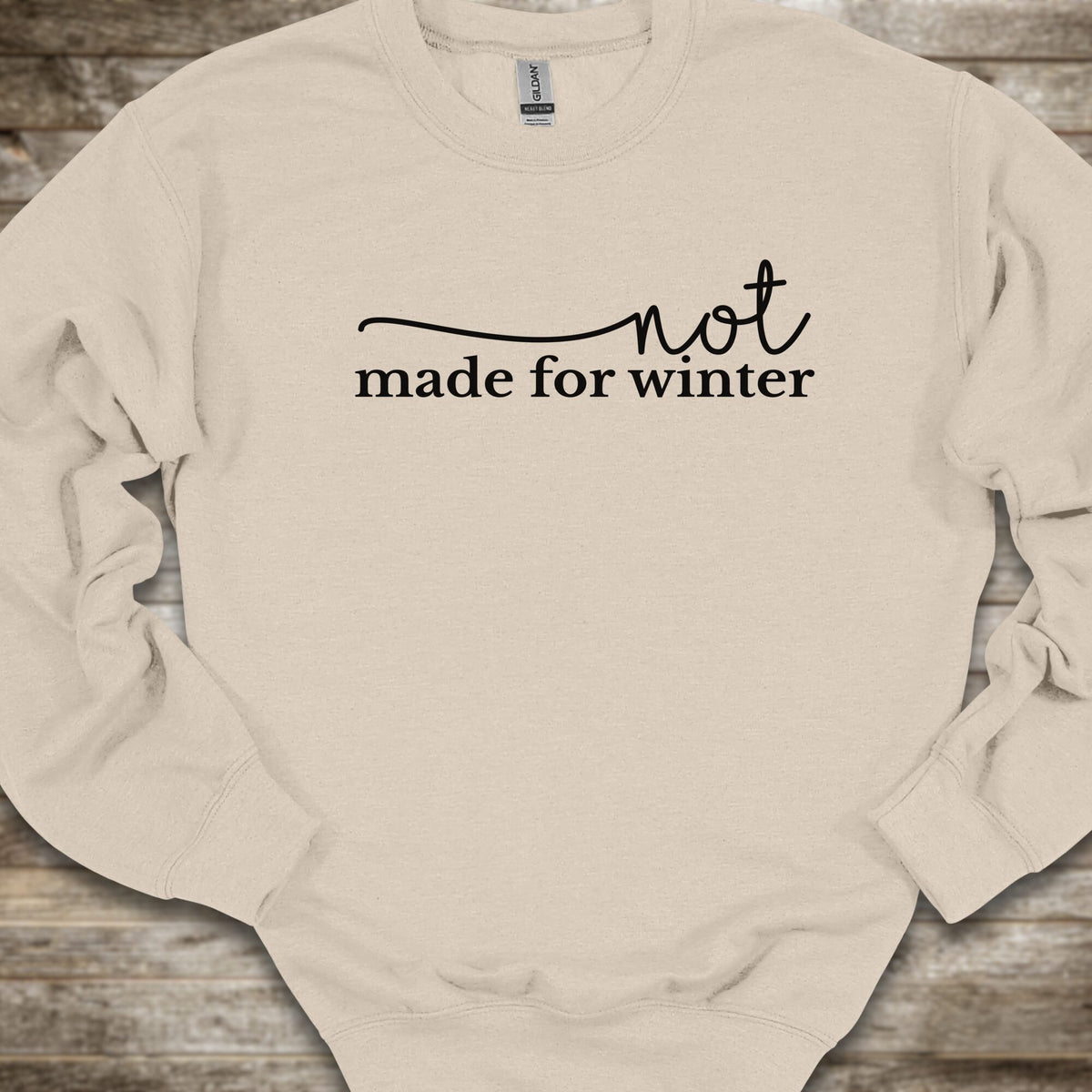 A cream-colored crewneck sweatshirt with the black printed text 'not made for winter' across the chest, laid flat on a rustic wooden background.