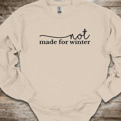 A cream-colored crewneck sweatshirt with the black printed text 'not made for winter' across the chest, laid flat on a rustic wooden background.