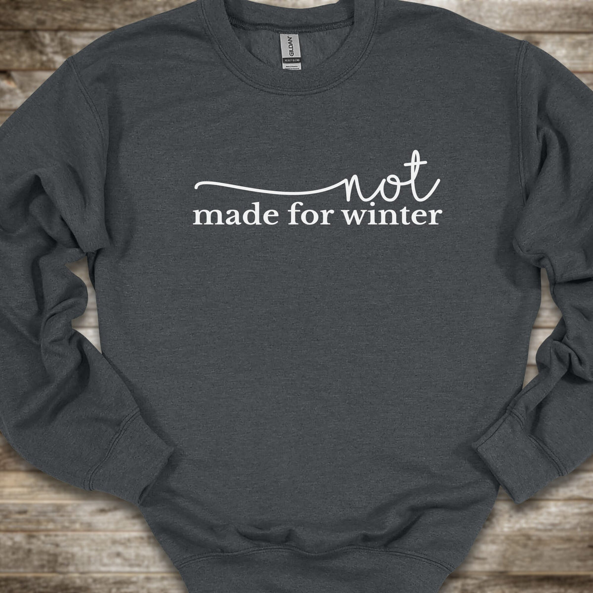 A dark gray crewneck sweatshirt with the white printed text 'not made for winter' across the chest, laid flat on a rustic wooden background.