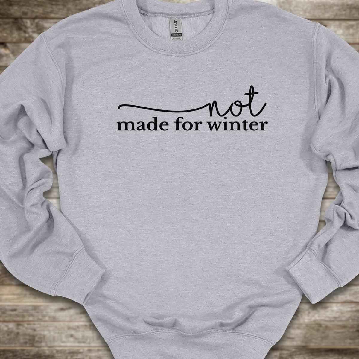 A light heather gray crewneck sweatshirt with the black printed text 'not made for winter' across the chest, laid flat on a rustic wooden background.